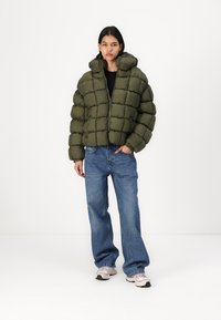 Grønn polstret kort pufferjakke med hette, med en quiltet design og frontglidelås, brukt over en svart topp og blå jeans.