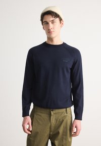 Mornarsko modra pletena pulover s dolgimi rokavi in krojnim ovratnikom. Ima diskreten logotip na prsih. V kombinaciji z olivno zelenimi hlačami.