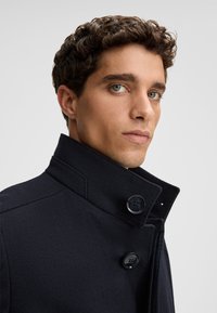 Marineblauwe wollen jas met een hoge kraag, voorzien van twee grote zwarte knopen en een gestructureerde stof. Close-up van de kraag en schouderdetail.