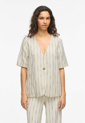 Kvinne med krøllete hår iført en kremfarget pinstripe kortermet blazer med én knapp og matchende bukser, stående foran en hvit bakgrunn.