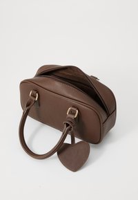 Sac à main en cuir marron aux bords arrondis, avec deux poignées et des accessoires en métal doré. Il possède une fermeture éclair et une étiquette en forme de cœur détachable.