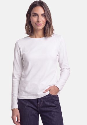 Femme aux cheveux bruns mi-longs, portant une chemise blanche à manches longues et un jean foncé, une main dans la poche, sur un fond uni.
