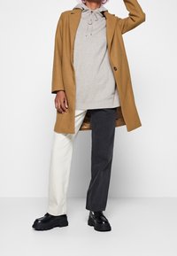 Brun kappa över grå hoodie, ljusa och mörka byxor. Svarta robusta skor. Enfärgade, minimalistisk design med en avslappnad och lager-på-lager-stil.