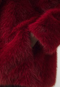 Rød faux fur kåpe med en fluffy tekstur, løs passform og vide ermer med en glatt innvendig fôr. Ingen synlige lukkinger eller maskinvare.