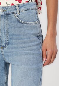 Jeans din denim albastru deschis cu talie înaltă, având cinci buzunare și detalii cu cusături aurii. Textură netedă cu ușoare decolorări.
