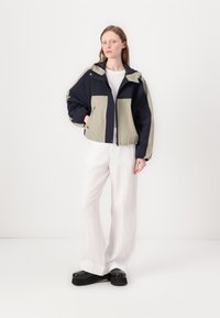 Veste en blocs de couleur bleu marine et vert clair, avec capuche, fermeture éclair à l'avant et poignets élastiques. Associée à un pantalon large blanc et des chaussures noires.