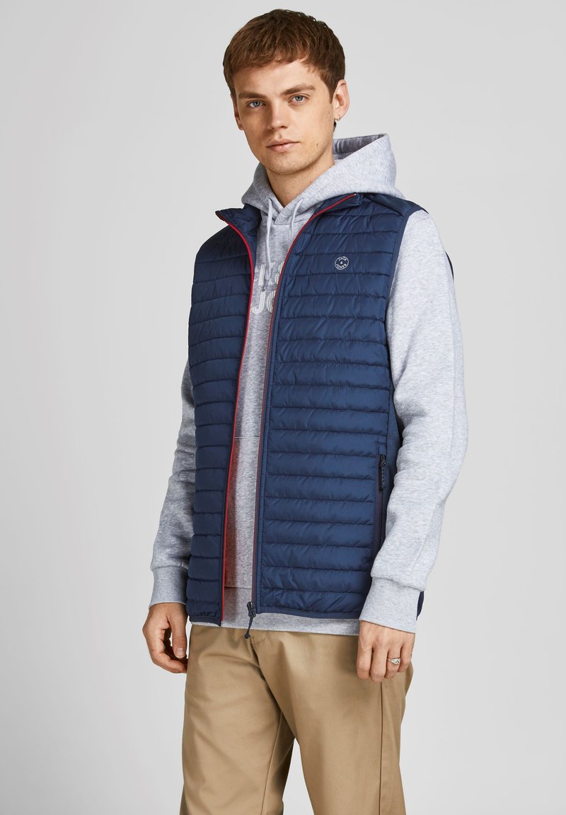 Marineblå quiltet vest med rød kant, lynlås-lommer og et logopatch. Båret over en grå hættetrøje og sammen med brune bukser. Letvægtsdesign.