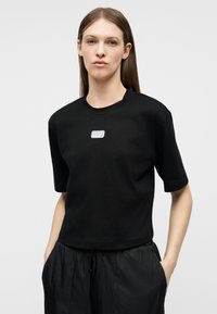 Γυναίκα με ίσια καστανά μαλλιά, που φοράει μαύρο μπλουζάκι με το λογότυπο EA7 και μαύρο παντελόνι, με τα χέρια στις τσέπες, στέκεται μπροστά σε απλό φόντο.