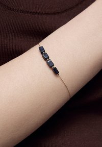 Bransoletka w kolorze złotym z pięcioma prostokątnymi ciemnoniebieskimi kamieniami, o gładkiej fakturze i minimalistycznym wzornictwie, noszona na jasnej karnacji.