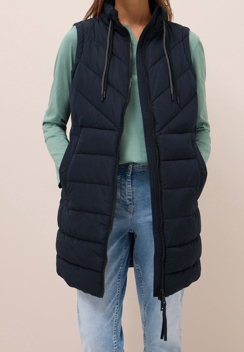 Femme portant une longue veste matelassée bleu marine avec capuche, ouverte à zip sur une chemise vert clair à manches longues et un jean bleu clair, mains dans les poches.