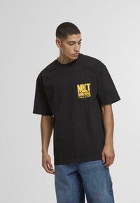 Czarny bawełniany t-shirt z okrągłym dekoltem, krótkimi rękawami i żółtą grafiką logo na lewej kieszeni na piersi. Noszony z niebieskimi dżinsami.