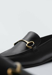 Schwarze Lederloafers mit einer abgerundeten Zehenpartie, die ein goldfarbenes metallisches Akzentdetail aufweisen. Glatte Textur mit einem dezenten Design.