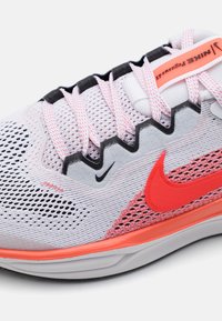 Chaussure de running Nike avec une tige en mesh grise et blanche, rehaussée par un logo swoosh rouge vif et un détail au talon, avec des lacets noirs et un motif texturé.