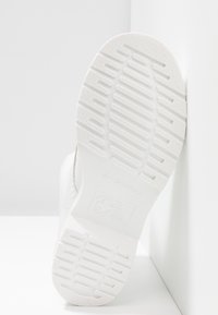 Suola di sneaker bianca con motivo in gomma texturizzata, marchio in rilievo e bordi laterali lisci, inclinata su uno sfondo chiaro.