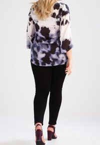 Top fluido multicolore con stampa bianca, nera e viola, abbinato a pantaloni neri aderenti e sandali con tacco viola scuro.