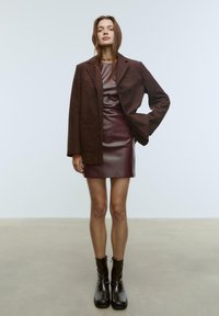 Brun mocka blazer över en bordeauxfärgad figurnära klänning med en glansig finish. Höga svarta stövlar kompletterar looken. Enkla bakgrund.