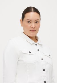 Chaqueta de mezclilla blanca con cuello, cierres de botón negros y dos bolsillos en el pecho. Textura suave y ajuste entallado.