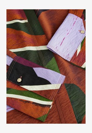 Multicolored fabric featuring bold patterns in rust, green, purple, and white. Includes detailed stitching and a decorative button accent.