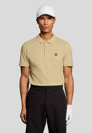 Man met beige poloshirt met geel vogellogo, zwarte broek, witte golfpet en witte handschoen aan de rechterhand, staand tegen een grijze achtergrond.