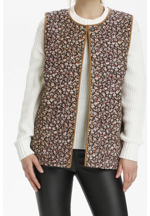 Osoba w brązowej, pikowanej kamizelce w kwiaty bez rękawów na białym dzianinowym swetrze i czarnych skórzanych spodniach, stojąca z rękami swobodnie opuszczonymi wzdłuż ciała.