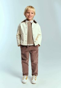 Jachetă albă din denim cu guler maro, pulover tricotat bej, cămașă cu carouri, pantaloni din corduroy maro și adidași albi cu accente galbene.