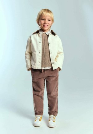 Veste en denim blanc avec col marron, pull en maille beige, chemise à carreaux, pantalon en velours côtelé marron et baskets blanches avec accents jaunes.