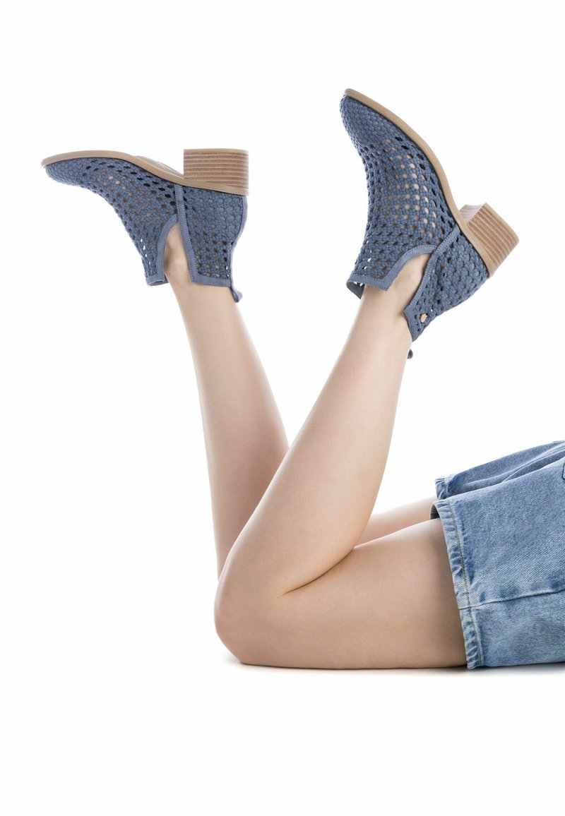 Botines tobilleros de hilo azul con un diseño perforado, cortes en los lados y un tacón cuadrado de color marrón claro. Se exhiben sobre un fondo blanco.