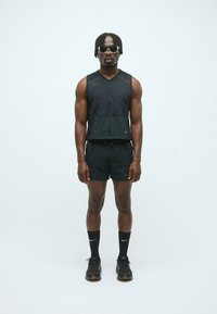 Schwarzes Sportoutfit bestehend aus einem ärmellosen Oberteil und Shorts, beide mit strukturiertem Finish. Zubehör umfasst schwarze Socken und Sneaker.
