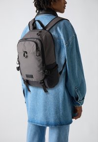 Sac à dos en tissu gris avec une surface texturée, poche zippée à l'avant et bretelles ajustables. Porté sur une veste en denim bleu clair.