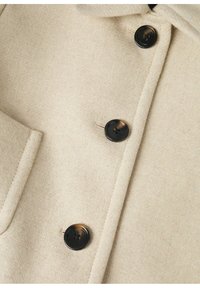 Chaqueta de mezcla de lana en color beige que presenta un acabado texturizado, cuatro botones negros y un detalle de bolsillo frontal. Diseño con cuello y solapas estructuradas.