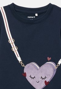 Granatowa koszula z nadrukowaną fioletową torebką w kształcie serca z zamkniętymi oczami, małymi czerwonymi serduszkami oraz paskiem w paski z zapięciami w kształcie serc.