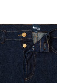 Donkerblauwe spijkerbroek met een Faguo-label, twee gouden knopen, gele stiksels en een zichtbare voorzak met een onderscheidende structuur.