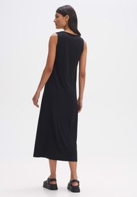 Zwarte mouwloze midi-jurk, gemaakt van zachte stof, heeft een relaxte pasvorm en valt tot halverwege de kuit. Gecombineerd met stevige zwarte sandalen.