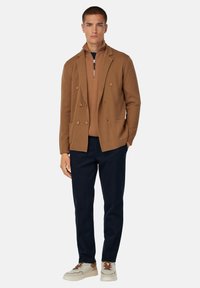 Chaqueta marrón de doble botonadura sobre un suéter marrón con cremallera, combinada con pantalones azul marino y zapatillas beige con detalles marrones.