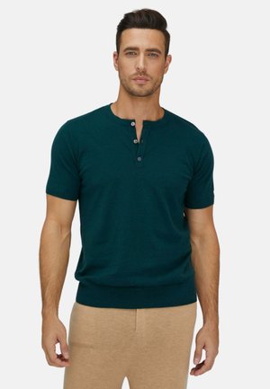 Mann mit kurzen dunklen Haaren, der ein dunkelgrünes Henley-Shirt mit kurzen Ärmeln und beigefarbene Hose vor schlichtem weißem Hintergrund trägt.