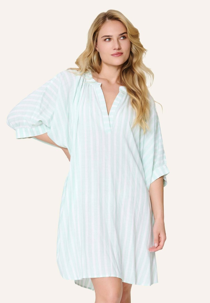 Femme blonde portant une robe ample à rayures verticales vert clair et blanches avec des manches trois-quarts, debout avec une main posée sur la hanche.
