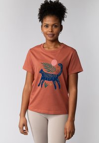 T-shirt en coton de couleur rouille avec un design stylisé de chat bleu, accompagné d'une feuille verte et d'un cercle rose. Manches courtes, col rond.