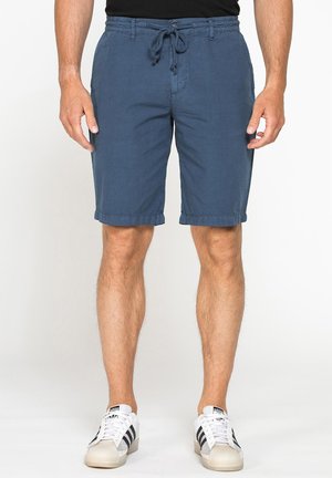 Uomo che indossa pantaloncini blu con coulisse e scarpe da ginnastica bianche, in piedi con le braccia rilassate lungo i fianchi su sfondo bianco.