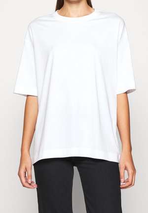 Persoon die een effen wit oversized T-shirt met korte mouwen en een zwarte broek draagt, staand tegen een neutrale achtergrond.