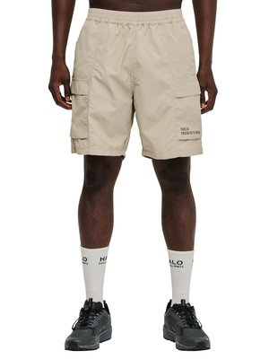 Beige cargoshorts met zijzakken gedragen door een staande persoon, gecombineerd met witte merk-sokken en zwarte sneakers.