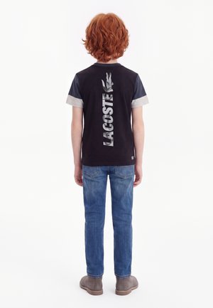 Enfant aux cheveux roux portant un t-shirt noir avec des manches grises et un texte vertical "LACOSTE" ainsi qu'un logo de crocodile dans le dos, jean bleu et chaussures marron.