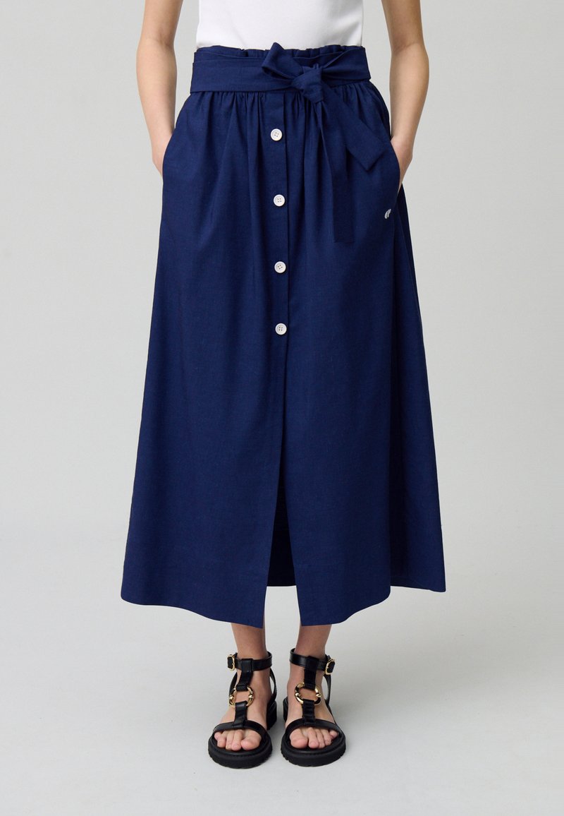 Jupe midi bleu marine avec une taille plissée, une ceinture en tissu et une fermeture à boutons sur le devant ; elle dispose de deux poches latérales et d'une texture lisse.