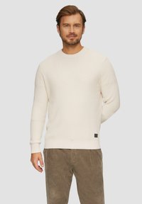 Crème waffle brei trui met geribde manchetten en zoom, met een klein zwart label detail, in combinatie met olijfgroene corduroy broek.