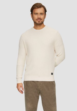 Crème waffle brei trui met geribde manchetten en zoom, met een klein zwart label detail, in combinatie met olijfgroene corduroy broek.