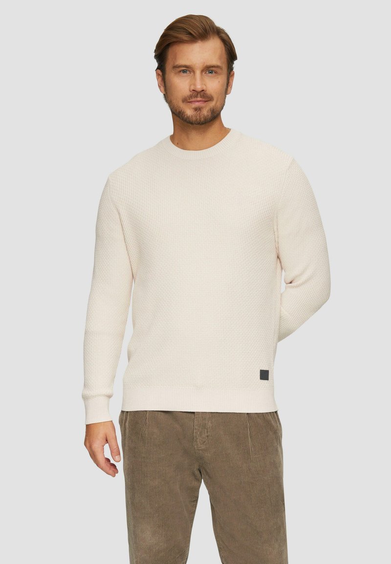 Crème waffle brei trui met geribde manchetten en zoom, met een klein zwart label detail, in combinatie met olijfgroene corduroy broek.