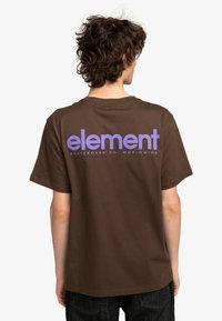 Camiseta de algodón marrón con gran texto en púrpura "element" y "SKATEBOARD CO. WORLDWIDE" debajo, con un corte relajado y mangas cortas.