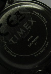 Czarny metaliczny tył zegarka z napisem "TIMEX", szczegółami dotyczącymi wodoszczelności, numerem modelu i znakiem CE w okrągłym wzorze.