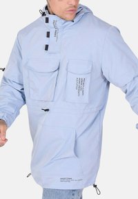 Pullover con cappuccio di colore azzurro chiaro, realizzato in tessuto a trama. Presenta toggles regolabili, quattro tasche e dettagli testuali stampati. Polsini elasticizzati.