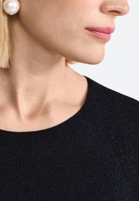 Black textured garment with a rounded neckline, paired with a large white pearl earring. Skin tone is neutral, lips have a soft pink shade.