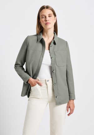 Veste verte à boutons, avec un col, une poche poitrine simple, des manches longues et un tissu lisse et texturé ; associée à un pantalon blanc.